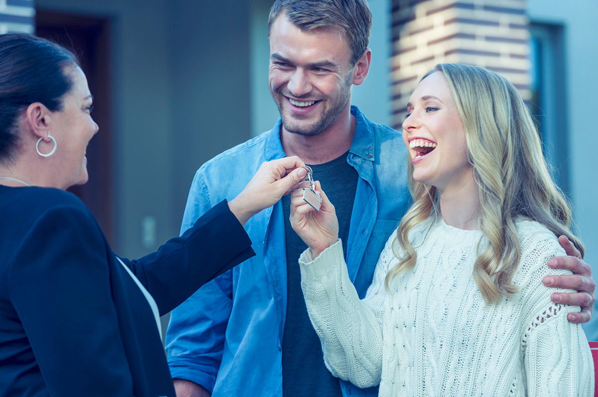 Couple standing in front of a new home with agent. They are standing next to a for sale sign with a sold sticker. They are buying or selling this real estate from a real estate agent. They are both wearing casual clothes and embracing. They are smiling and he has a beard. The house is new and contemporary with a brick facade.