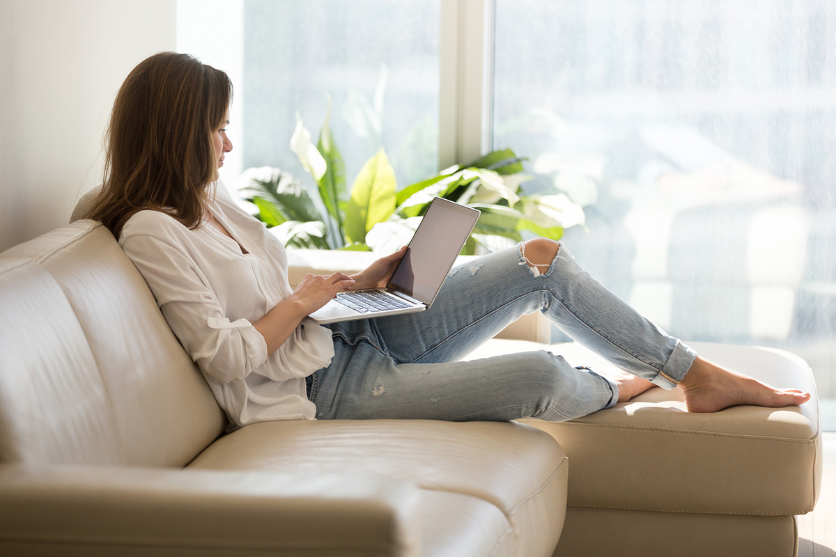 Calm female working at laptop sitting on cozy sofa, happy girl browsing internet or shopping online during sunny weekend at home, woman spending day off at home, relaxing on couch with computer