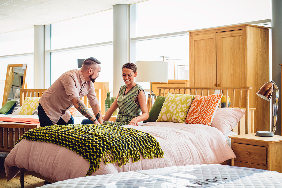 A middle aged couple are shopping in a furniture store for a new bedroom, the customers are both sat on the bed to see how comfortable it is, trying it out.